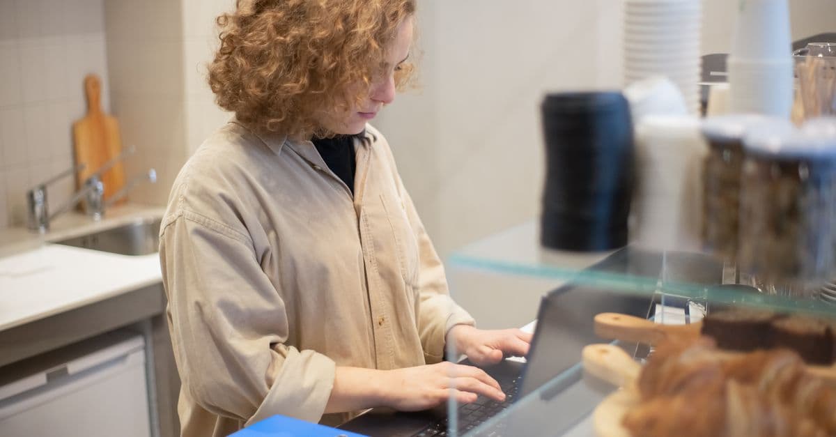 A cafe owner working on a laptop behind the counter, beside the pastry display