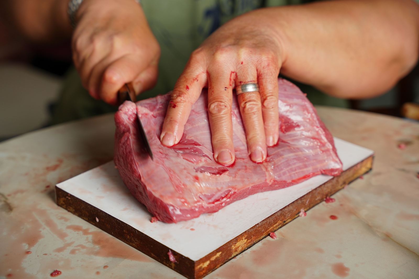 A detailed image of hands with a knife cutting a piece of raw meat on a cutting board indoors